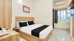 A hotel room with a wooden bed, a desk with a chair, a TV, and a window with blinds. - RG Suites HSR (Bengaluru)