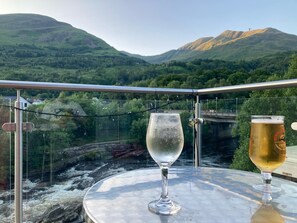 Een tafel met twee bierglazen en een wijnglas, met uitzicht op een rivier en bergen.