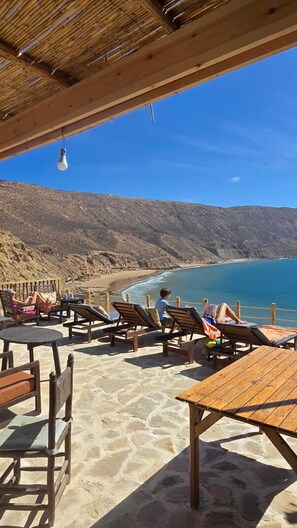 An outdoor seating area with wooden tables and chairs, overlooking a beach with clear blue waters and rocky hills in the background. - Aloha imsouane chambres (Imsouane)