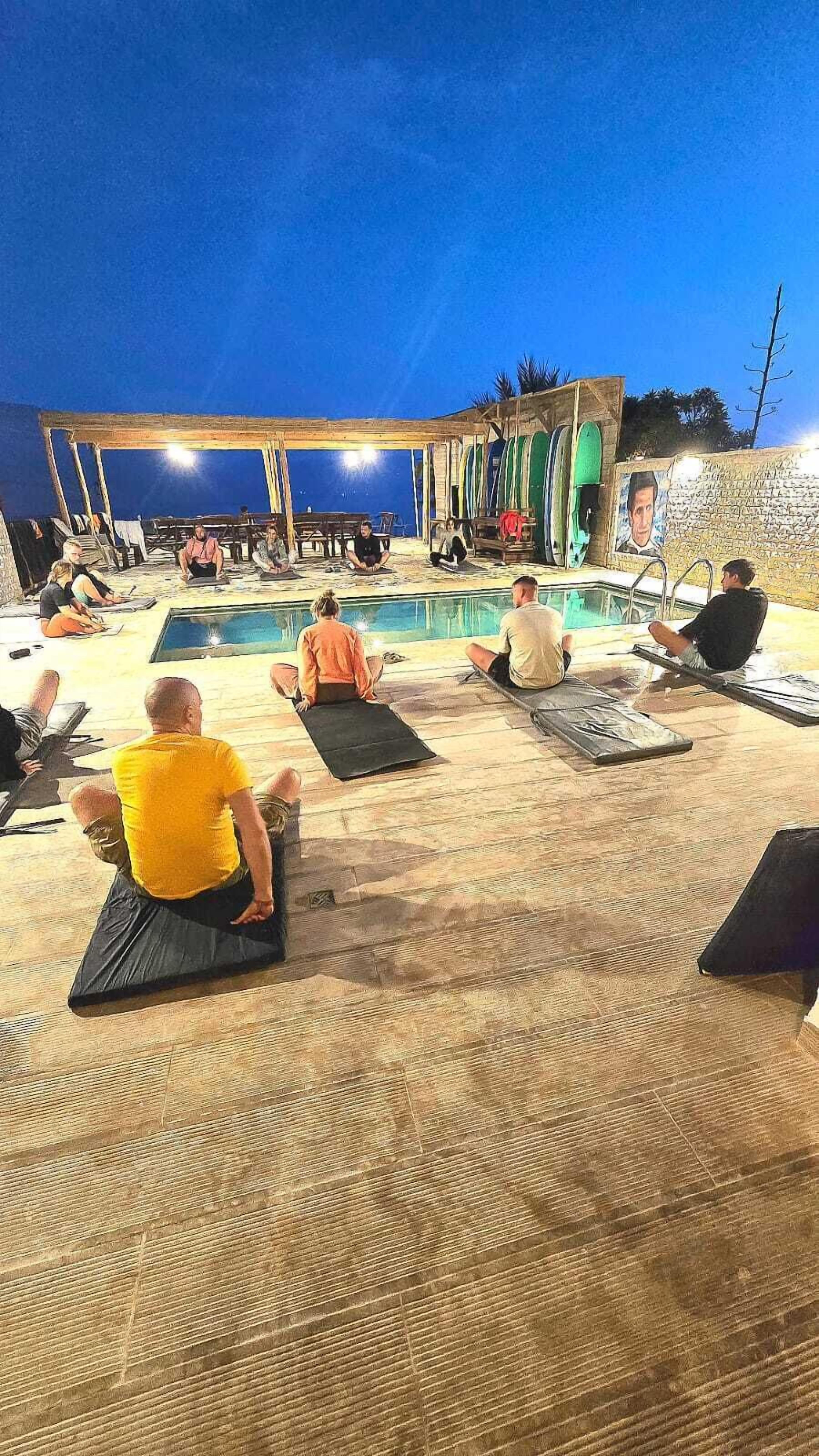 A group of people sitting on mats outdoors at dusk, with a pool and surfboards in the background.