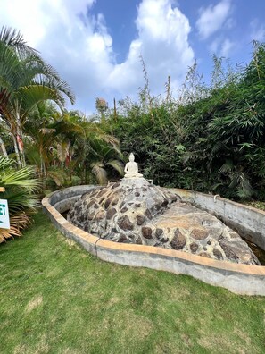 A stone fountain with a Buddha statue in the center, surrounded by lush greenery and a clear sky. - Plumeria Resort (Somvarpet)