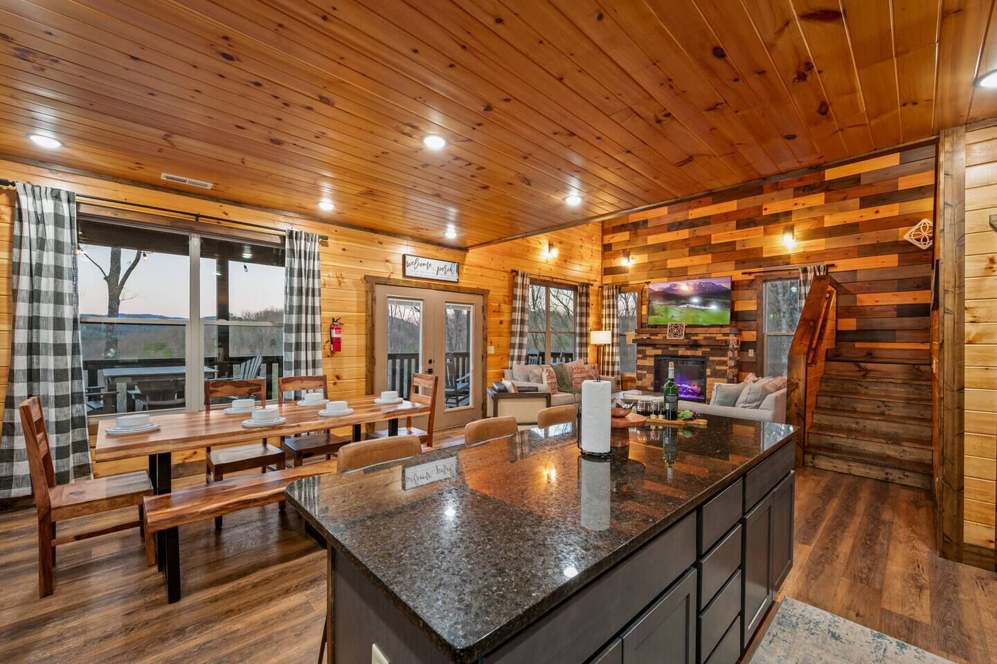 Un intérieur de chalet chaleureux, avec un revêtement mural en bois, un coin repas avec des bancs, une cuisine dotée d’un îlot central et un espace de vie agrémenté d’une cheminée.