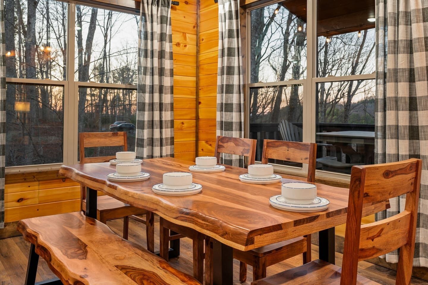A wooden dining table with white plates and chairs, set in a cabin with large windows and plaid curtains.