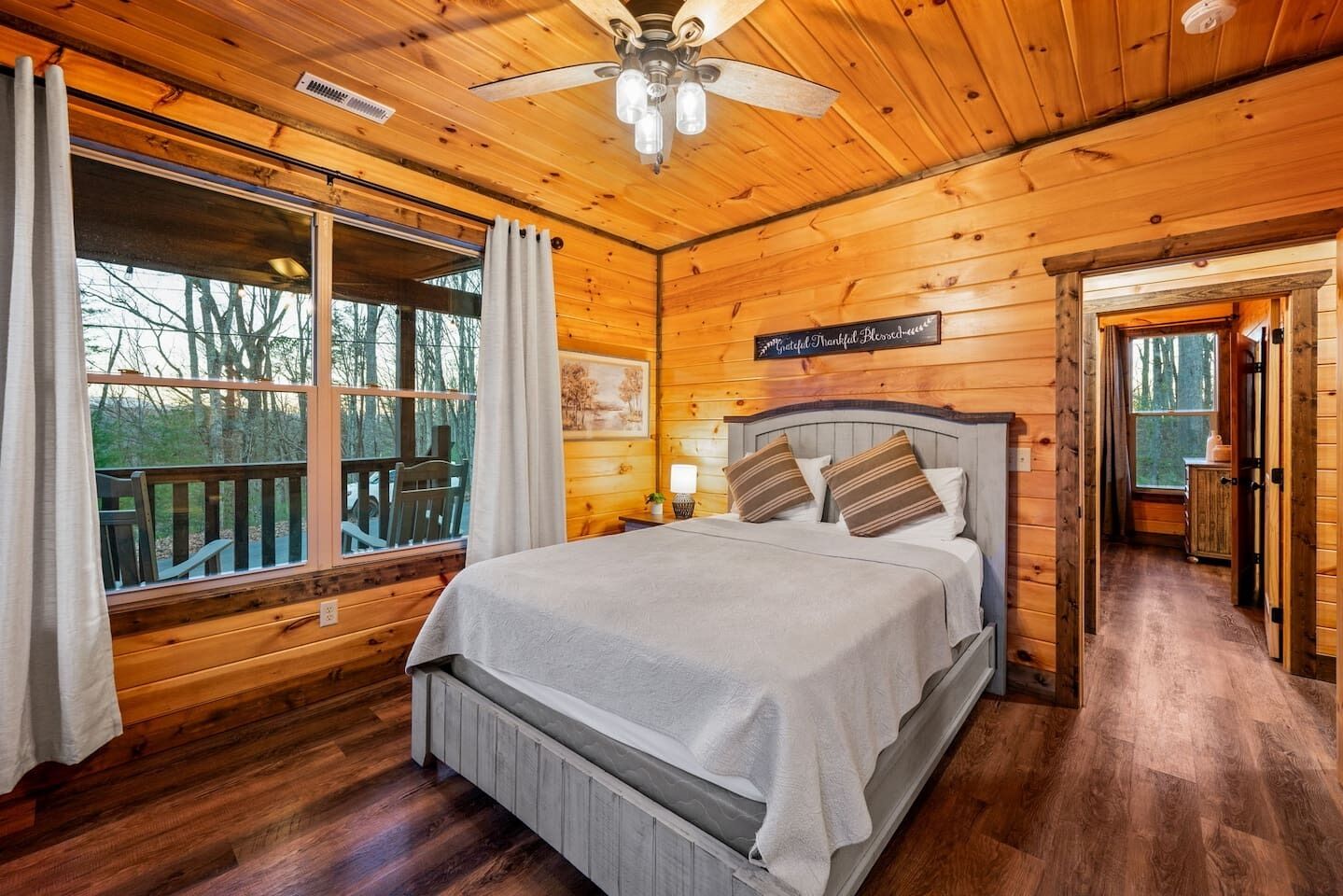 Quarto de cabana de madeira com cama, ventilador de teto e janela com vista para um deck.