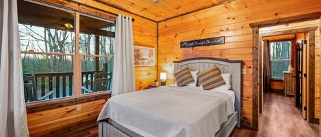 Quarto de cabana de madeira com cama, ventilador de teto e janela com vista para um deck.