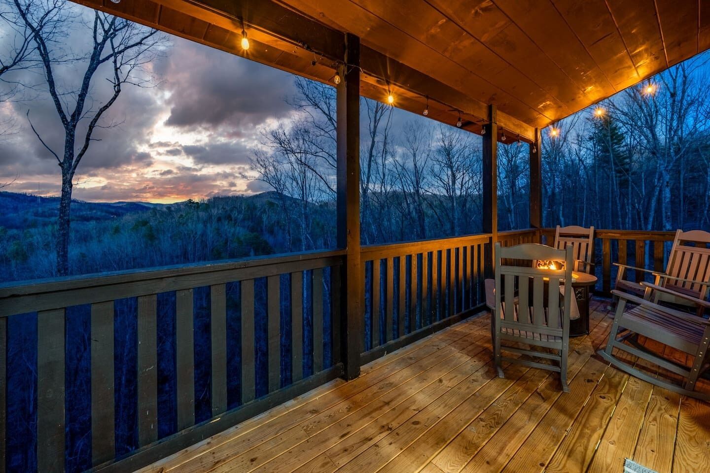 A wooden deck with a fire pit and chairs, overlooking a forested landscape.