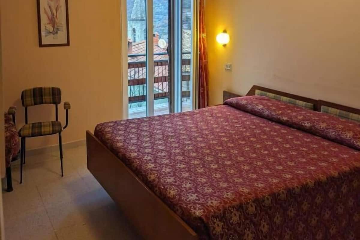 Une chambre à coucher avec un lit, une chaise, un tableau et une vue sur un immeuble, visible à travers une porte vitrée.