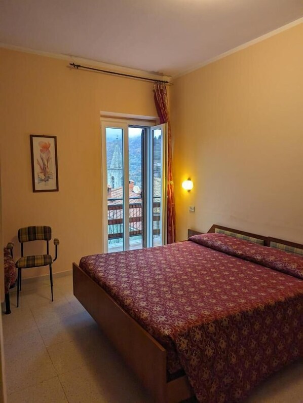 A bedroom with a bed, a chair, a painting, and a view of a building through a glass door.