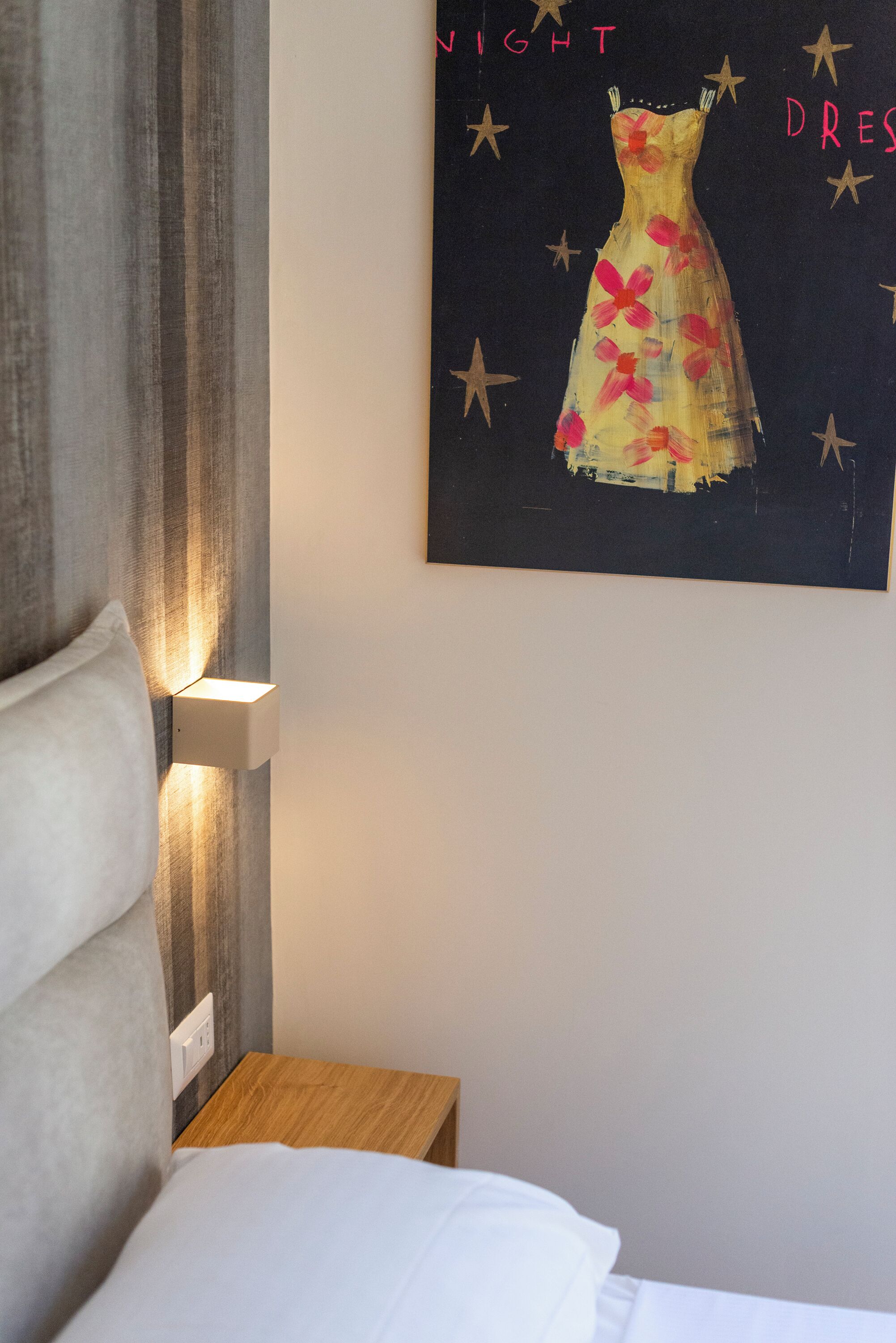 A bedroom with a bed, a nightstand, a wall-mounted light, and a painting of a dress on the wall.