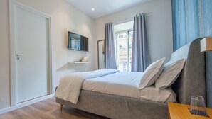 A bedroom with a bed, a TV mounted on the wall, a window with curtains, and a wooden floor. - Design 18 Charming Rooms (Rome)