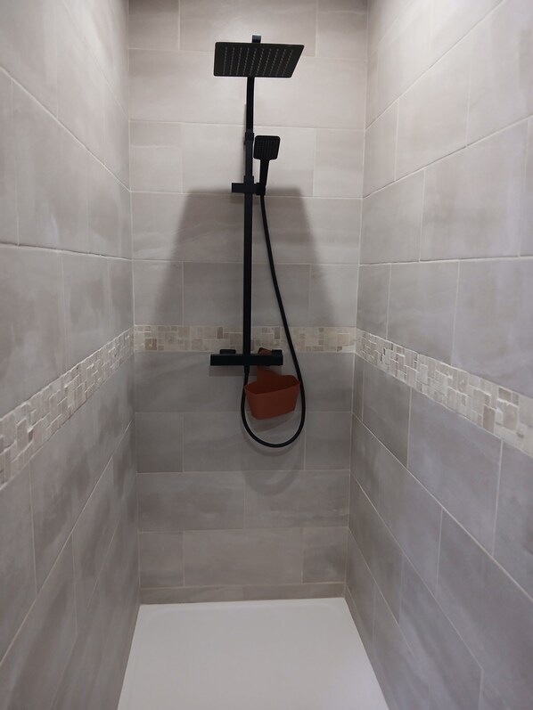 A shower area with a handheld showerhead, a handheld scrubber, and a red bucket.