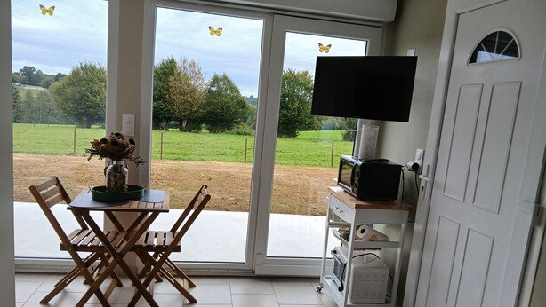 A room with a dining table, chairs, a bed, a TV on a stand, and a view of a green landscape through large windows. - La Chambrée (Leschelle)