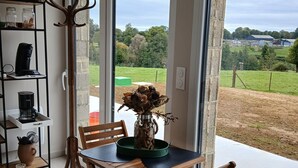 A dining area with a table and chairs, a coffee machine, a vase with dried flowers, and a view of a green field through large windows. - La Chambrée (Leschelle)
