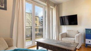 Una habitación con cama, una mesa de madera, un sofá, un televisor montado en la pared y una puerta corrediza de cristal que da a un balcón exterior.