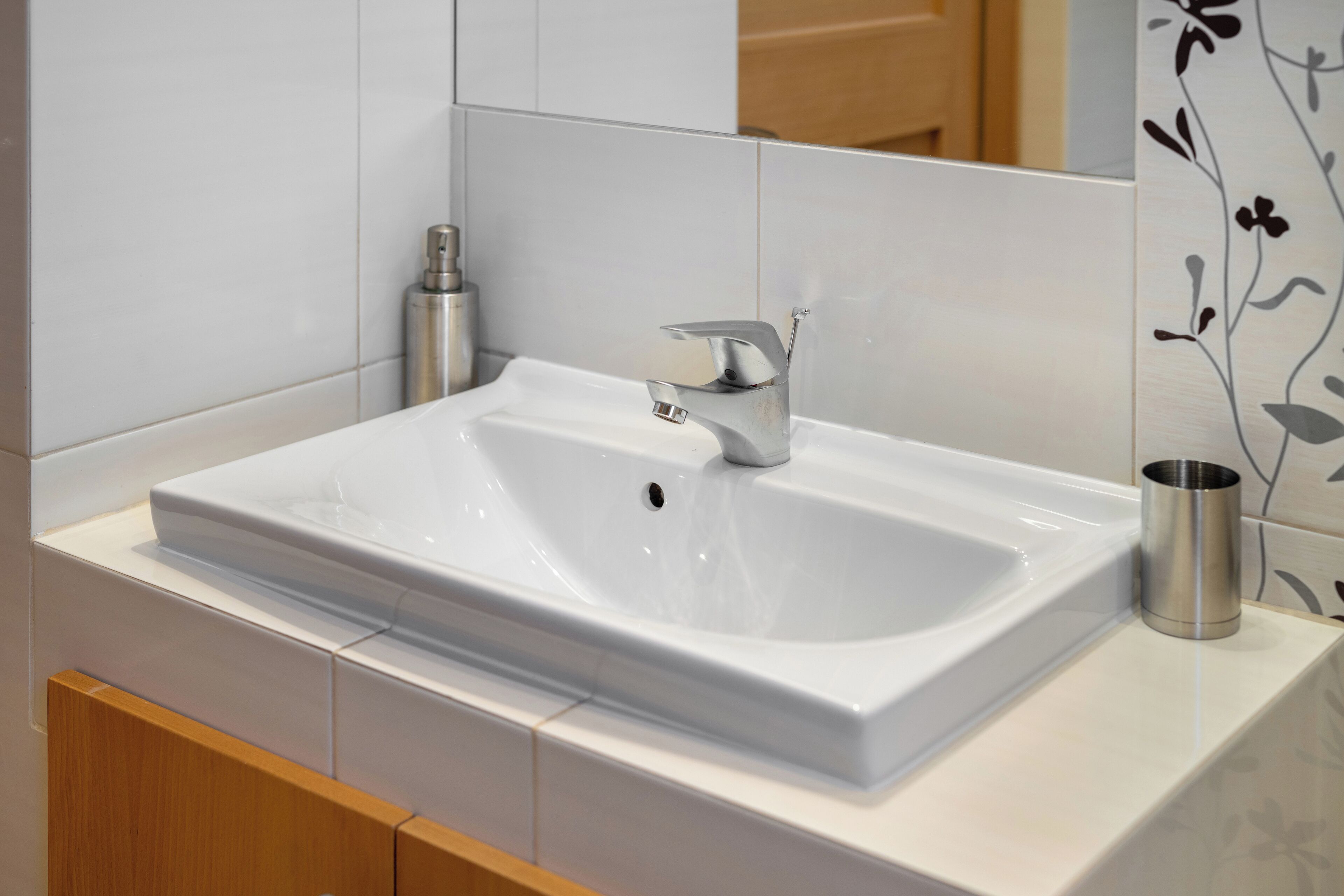 A modern bathroom with a white sink, a silver faucet, and a stainless steel cup on the counter.