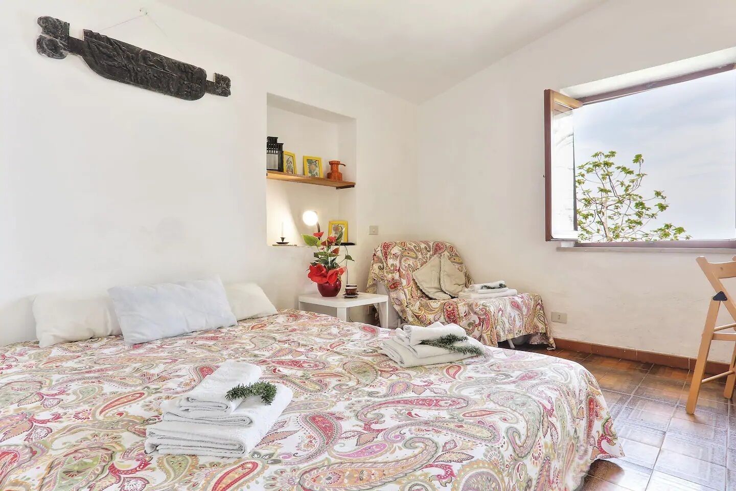 A bedroom with a large bed, a chair, a small table, and a window with a view of trees.