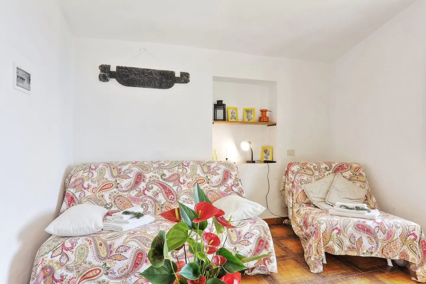 Une pièce comprenant un canapé et un fauteuil, tous deux recouverts d’un tissu à motifs cachemire. On y trouve une étagère avec des photos encadrées et une petite table avec une lampe.