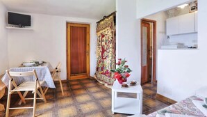 Una habitación pequeña y bien iluminada con una puerta de madera, un televisor montado en la pared, una mesa de comedor con sillas, y una pequeña mesa blanca con un jarrón de flores.