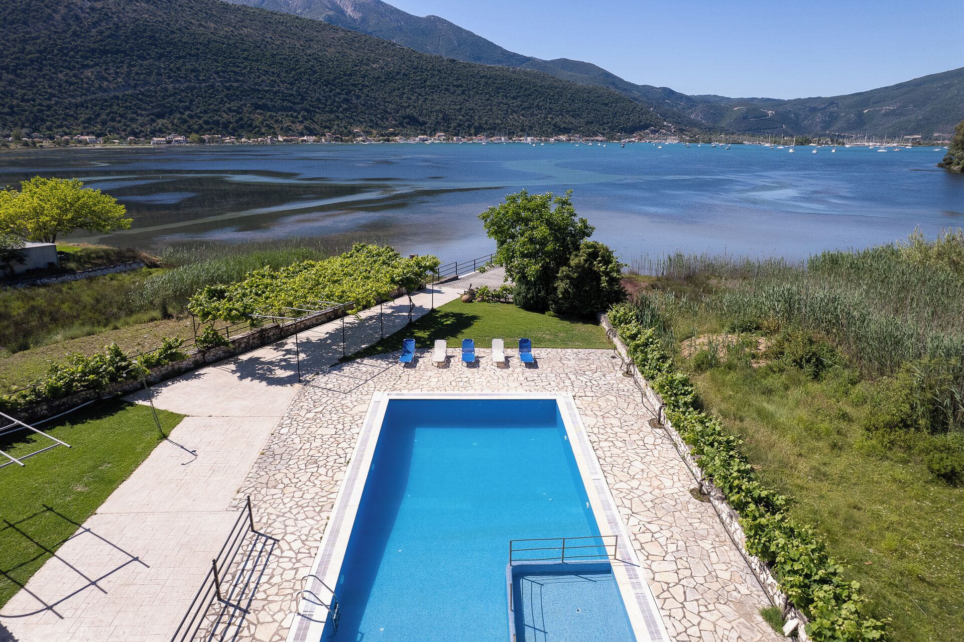 A rectangular swimming pool with a stone border, surrounded by a stone deck and a grassy area with a few trees.