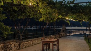 Un espace de restauration en plein air, avec un mur en pierre et une table entourée de chaises, offrant une vue sur une étendue d’eau et sur le paysage urbain au loin.