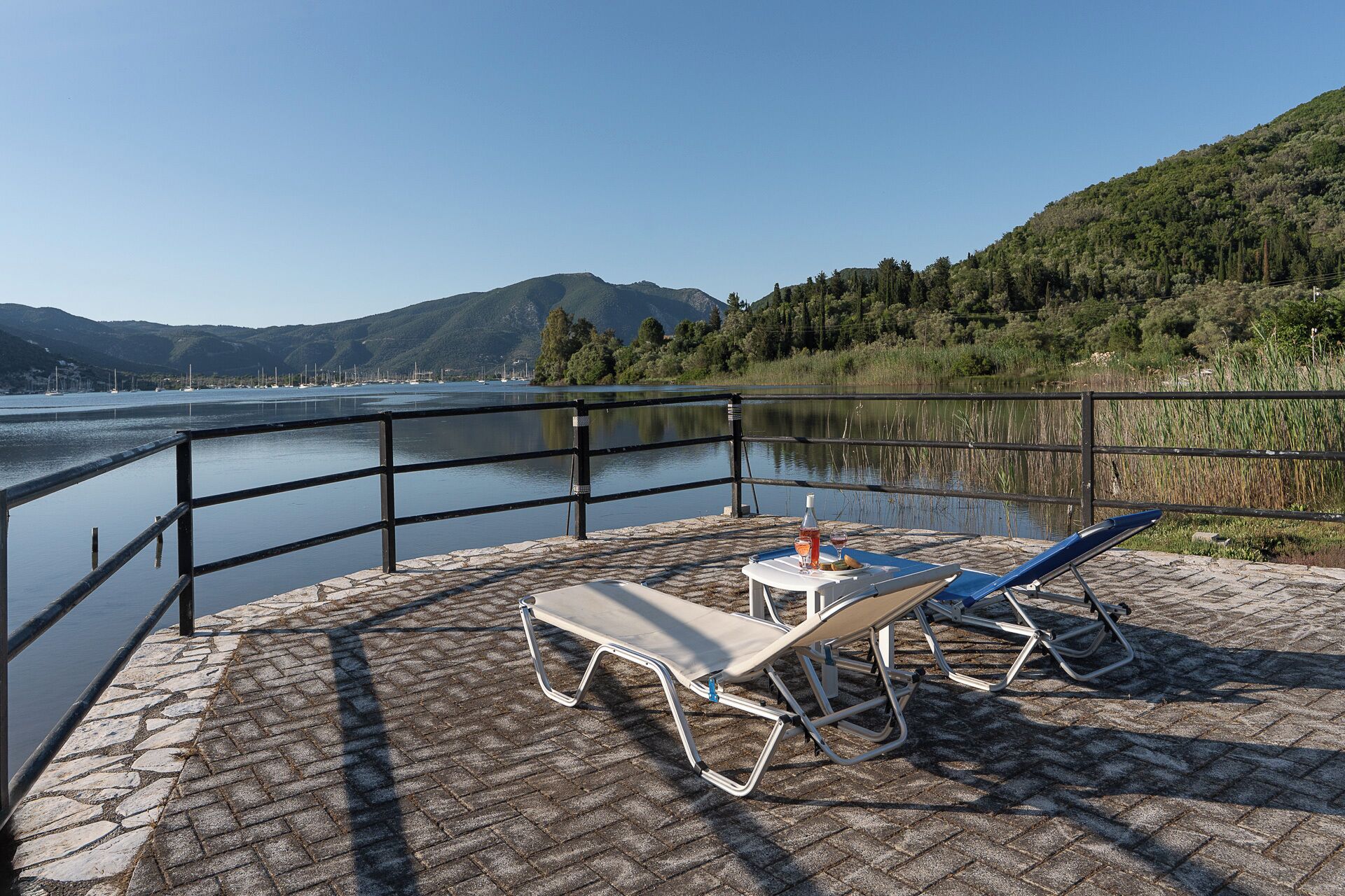 A lakeside patio with two lounge chairs, a table with drinks, and a view of boats and mountains.