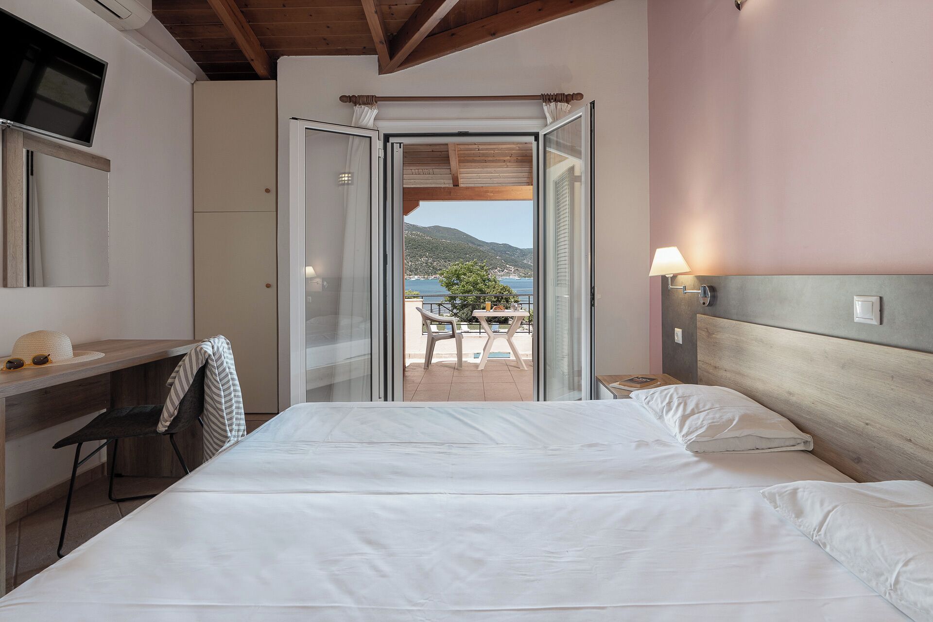 A bedroom with a bed, a desk, and a view of a balcony with a table and chairs.