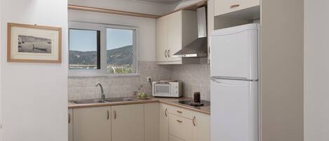 Cocina pequeña con armarios blancos, microondas integrado y refrigerador. Una ventana ofrece vistas a un paisaje costero.