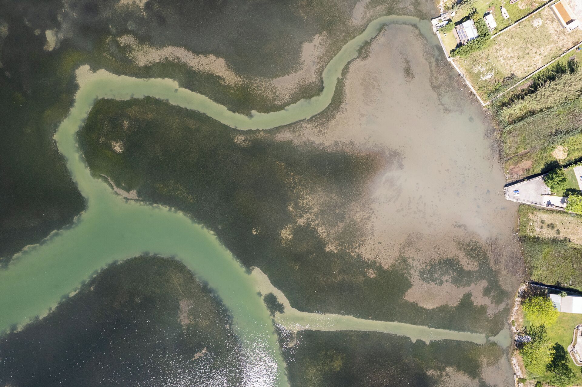 Vista aérea de una zona costera con una masa de agua verdosa, áreas arenosas y un pequeño conjunto de edificios.