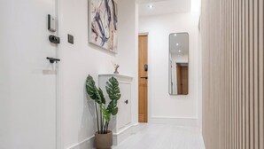 A hallway with a white door, a potted plant, a wooden cabinet, and a mirror on the wall. - Cosy Nook | Kitchen w/ Washer & SofaBed | Free Parking (Merseyside)