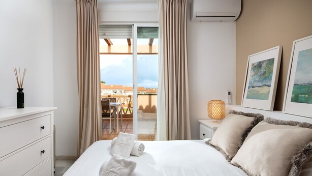A hotel room with a bed, a nightstand, a lamp, and a view of the outside.