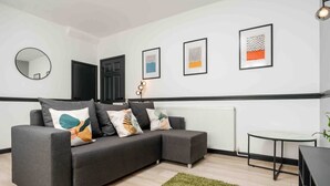 A modern living room with a grey sectional sofa, a round mirror, and framed artwork on the wall. - 10Min Walk to Stadium | SofaBed | W/M & Dishwasher (Anfield)