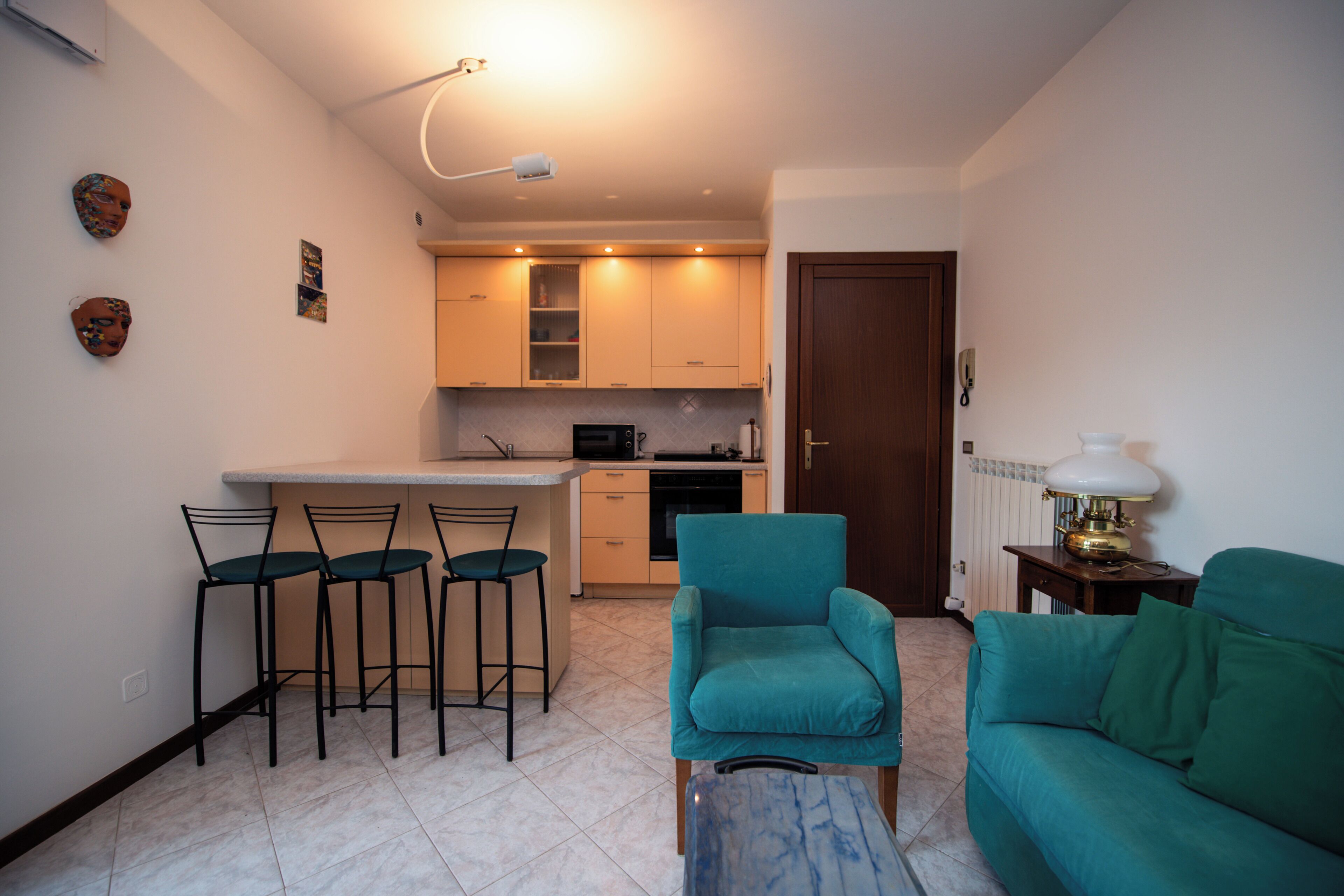 A compact living space with a kitchenette, a dining area with bar stools, a teal armchair, and a sofa.