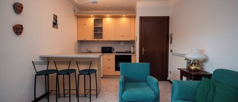 A compact living space with a kitchenette, a dining area with bar stools, a teal armchair, and a sofa.