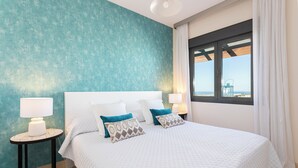 Een slaapkamer met een groot bed, twee bedlampen, uitzicht op zee door een raam, en een wand in turquoise.