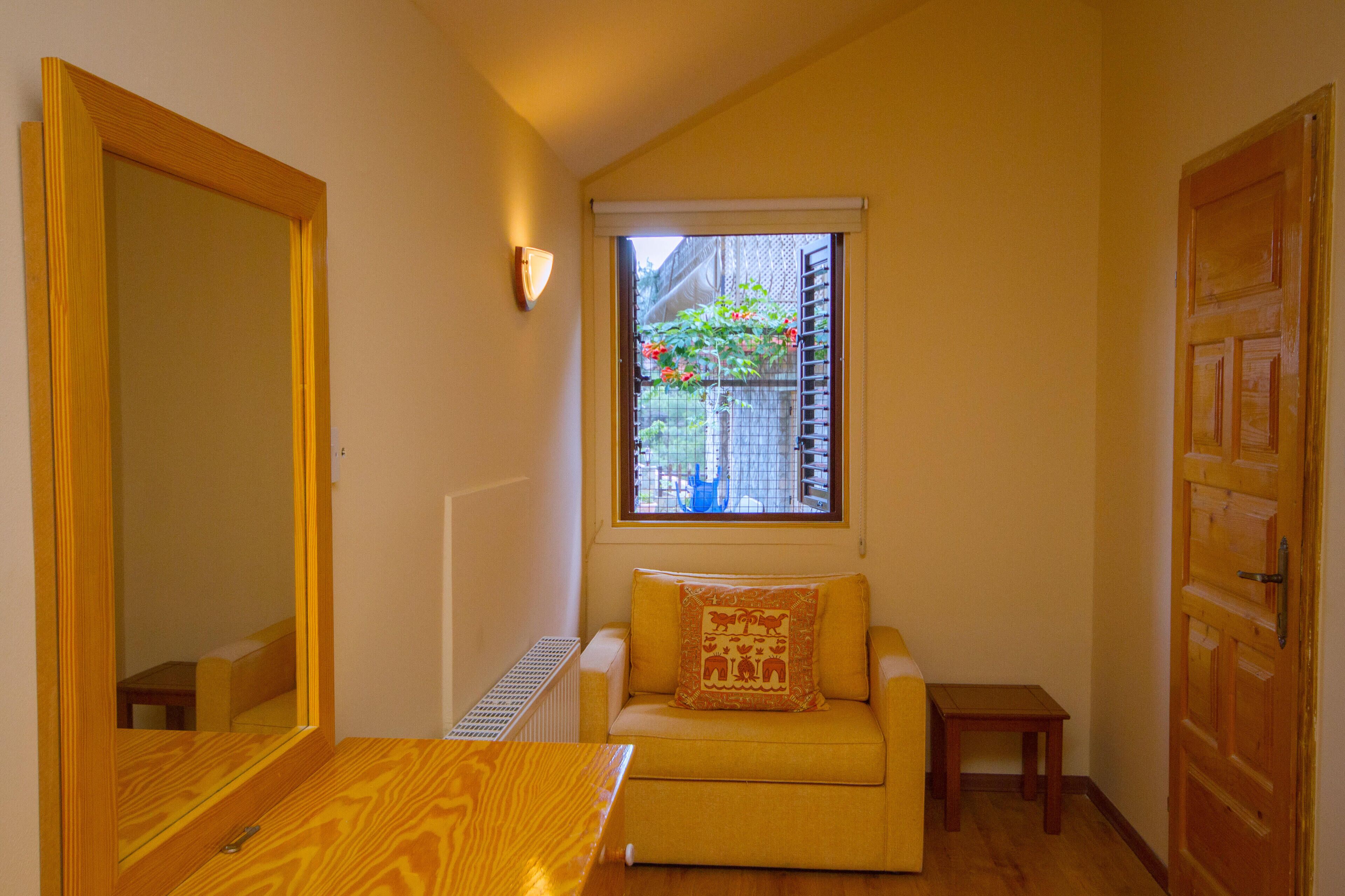 Una habitación pequeña con un sofá amarillo, una mesa de madera, un ventanal con vistas a un jardín y una puerta de madera.