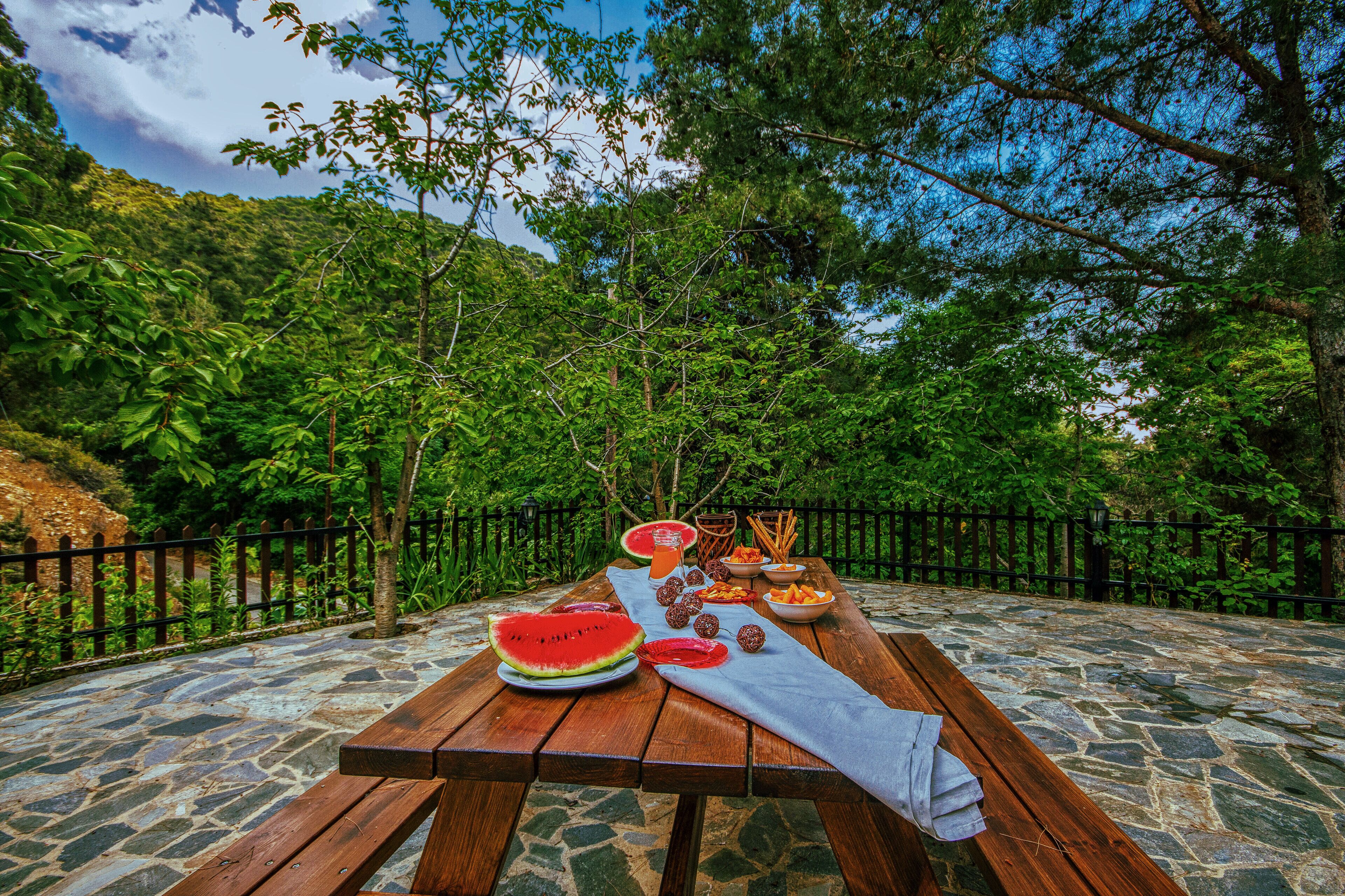 A picnic table set on a stone-paved area with a wooden bench, surrounded by lush greenery and a metal fence.