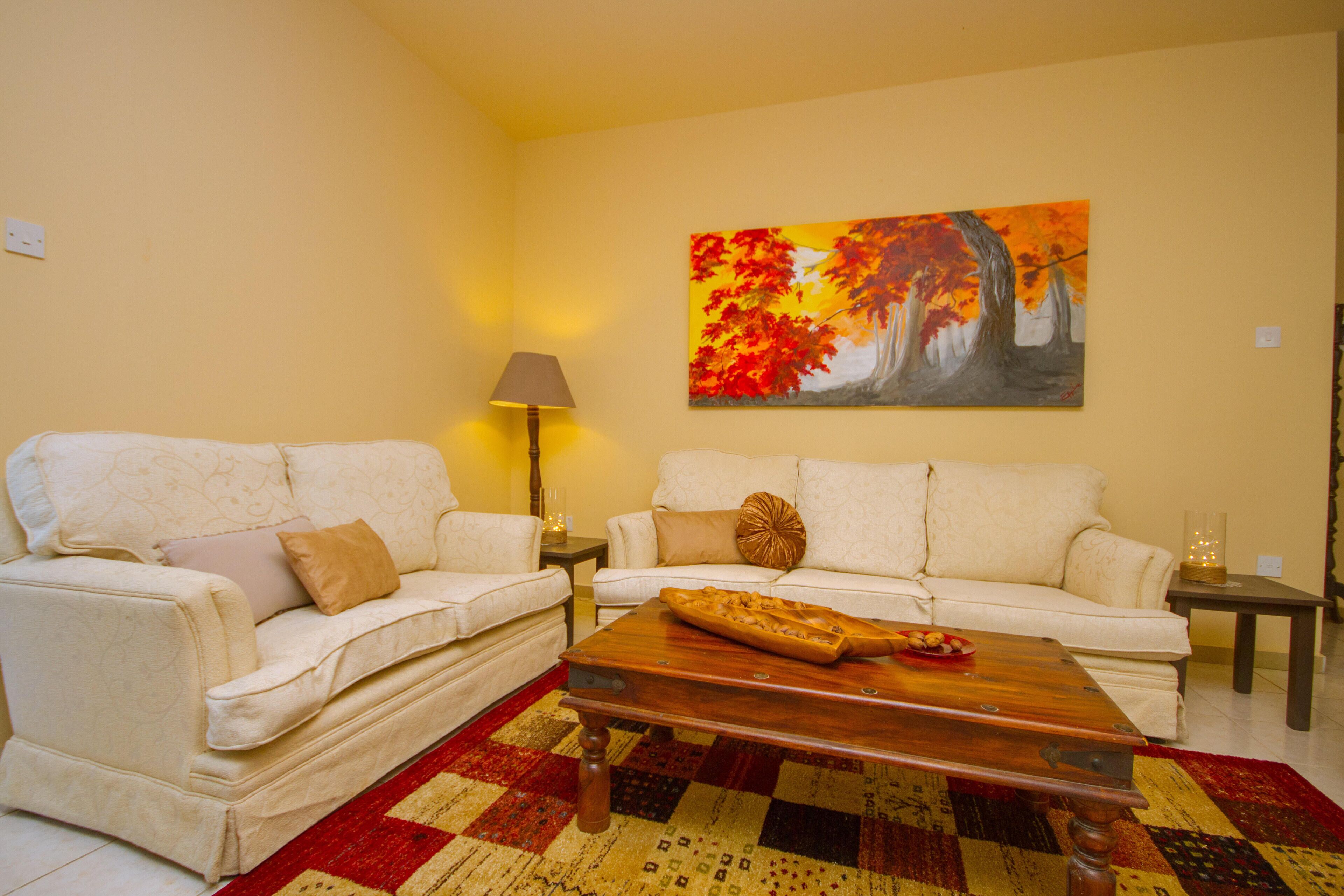Sala de estar con dos sofás blancos, una mesa de centro de madera, un tapete estampado en rojo y amarillo, y un cuadro de árboles otoñales en la pared.