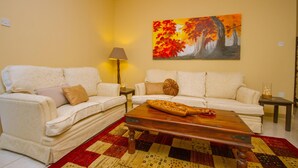 Un salon avec deux canapés blancs, une table basse en bois, un tapis aux motifs rouges et jaunes, et un tableau représentant des arbres d’automne accroché au mur.