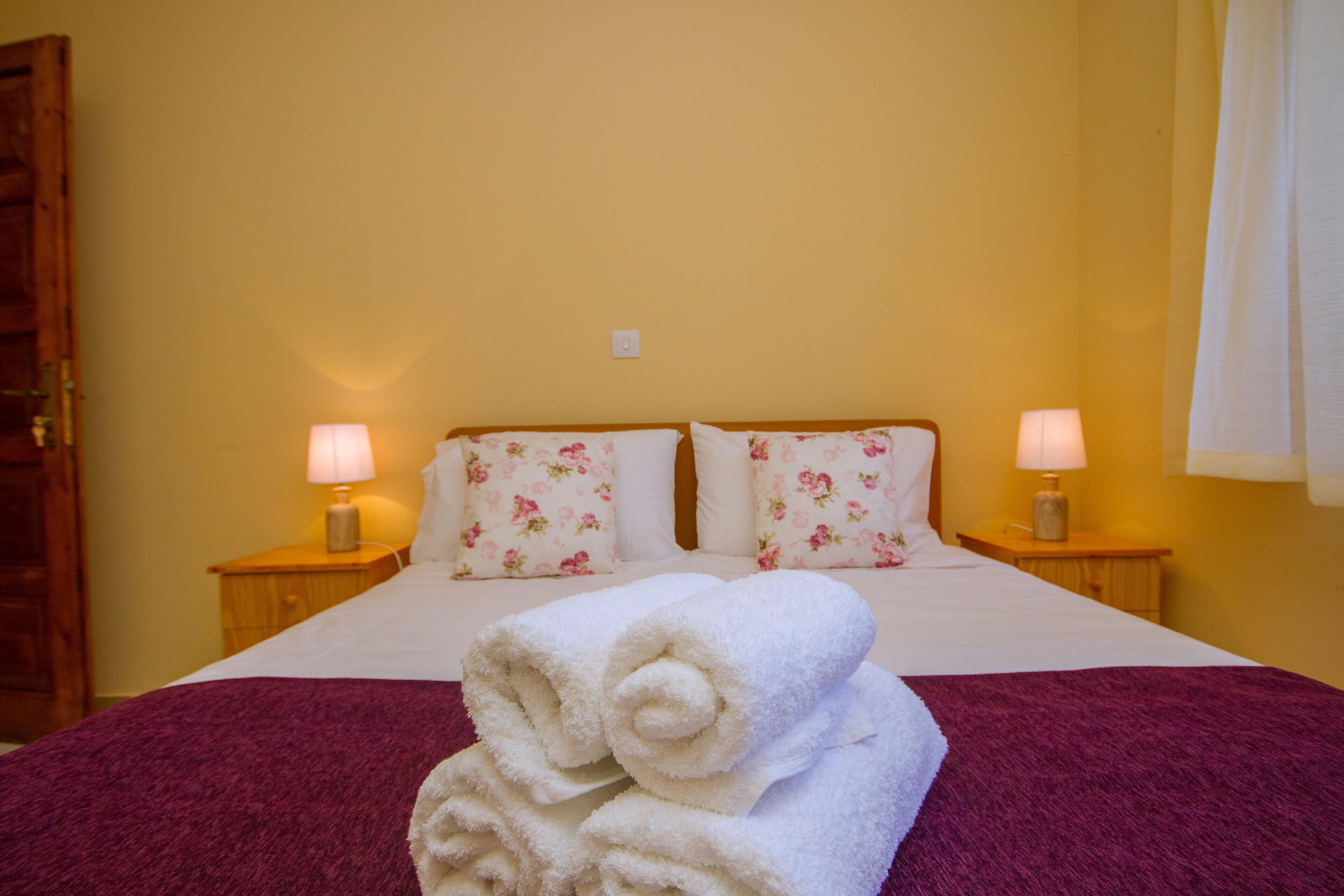 Habitación de hotel con cama, almohadones florales, dos lámparas de noche, una toalla blanca enrollada y una mesita de noche de madera.