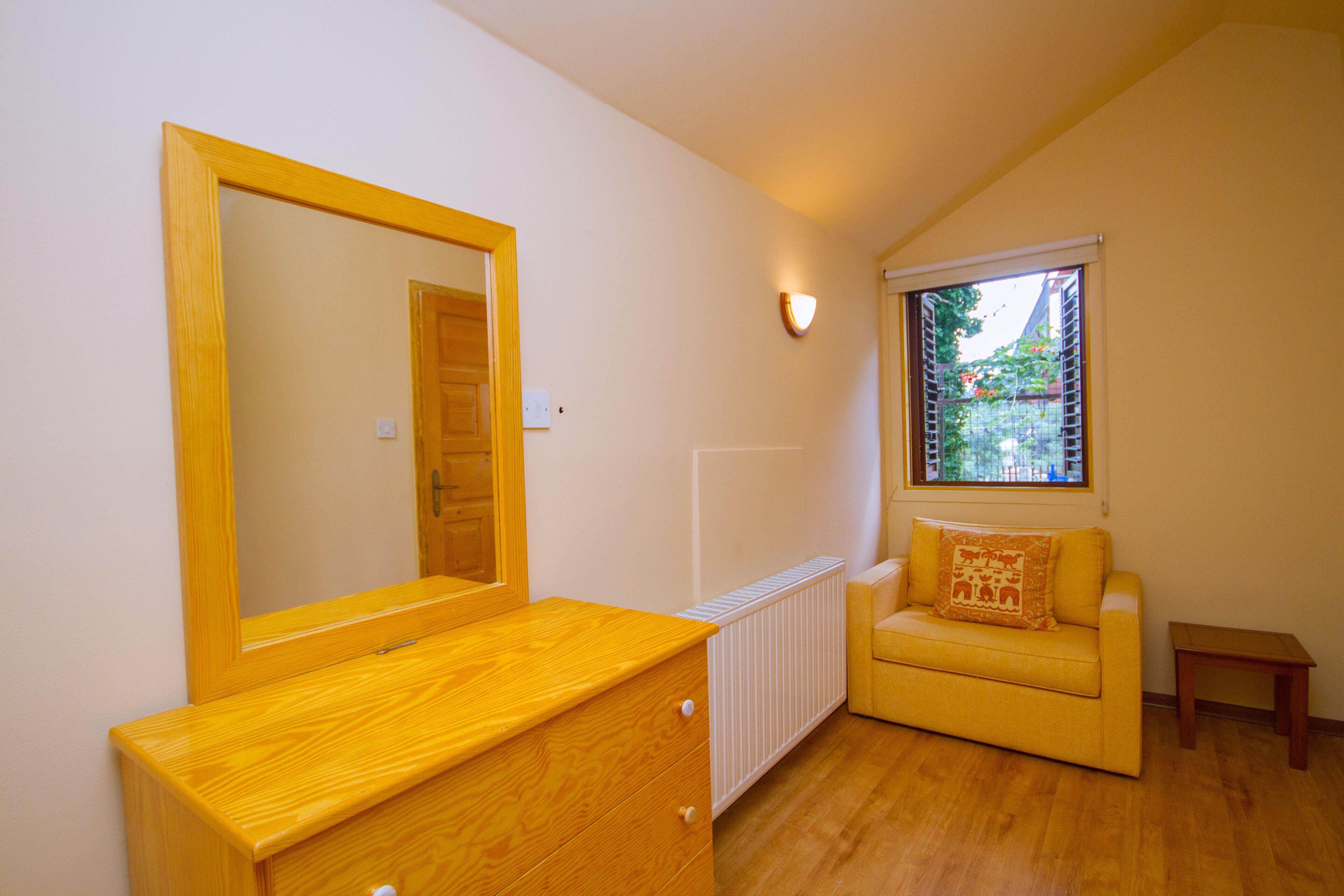 Una habitación con tocador de madera, espejo, radiador, sofá amarillo, una mesita y ventana con vistas a follaje.