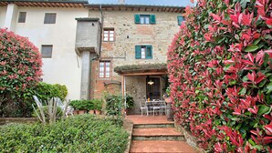 A stone building with a patio area featuring a table and chairs, surrounded by lush greenery and a brick pathway. - Francigena Pergola Apartment Shared Pool (Gambassi Terme)
