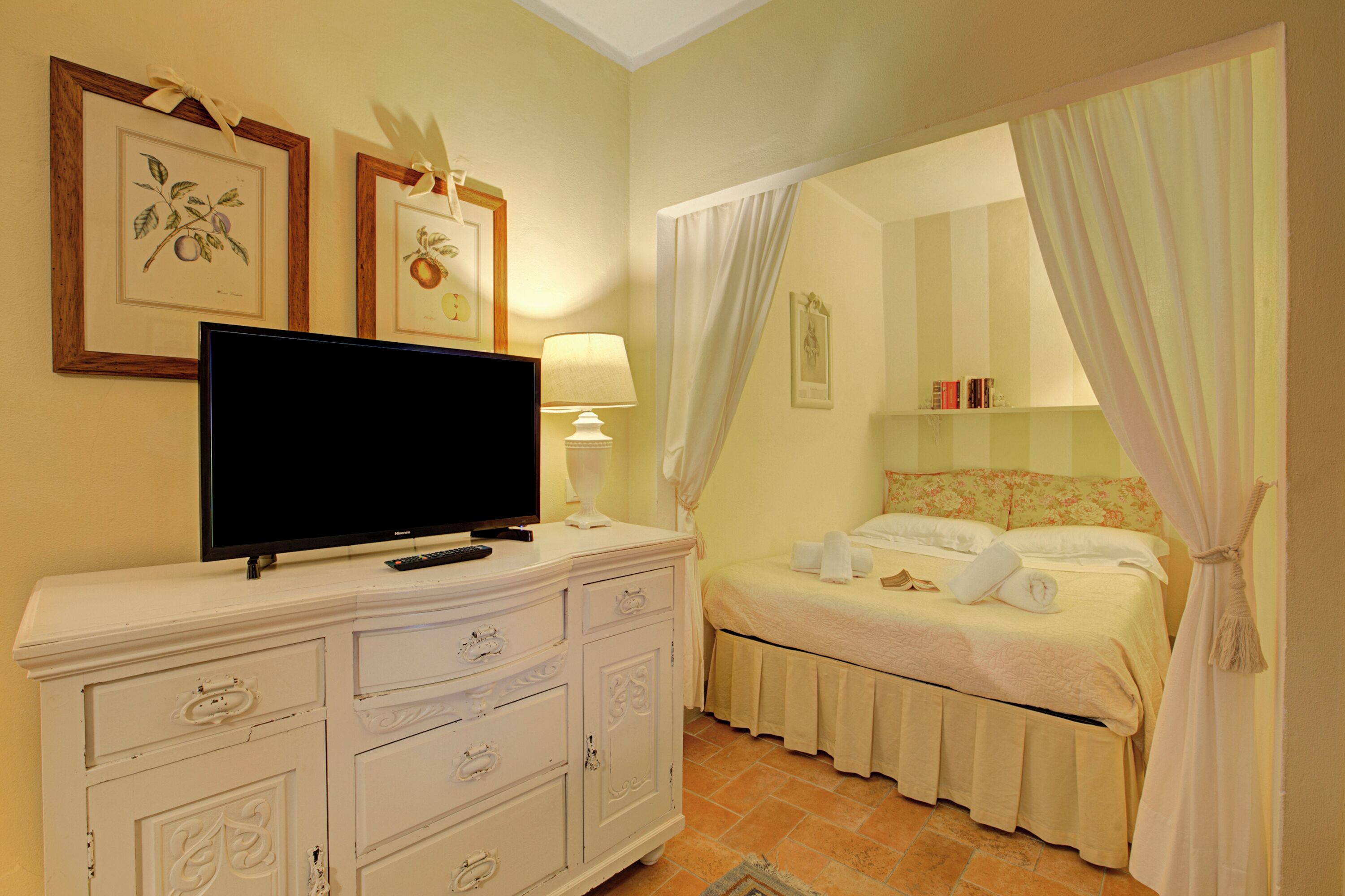 A bedroom with a television on a dresser, a bed with a canopy, and framed botanical prints on the wall.