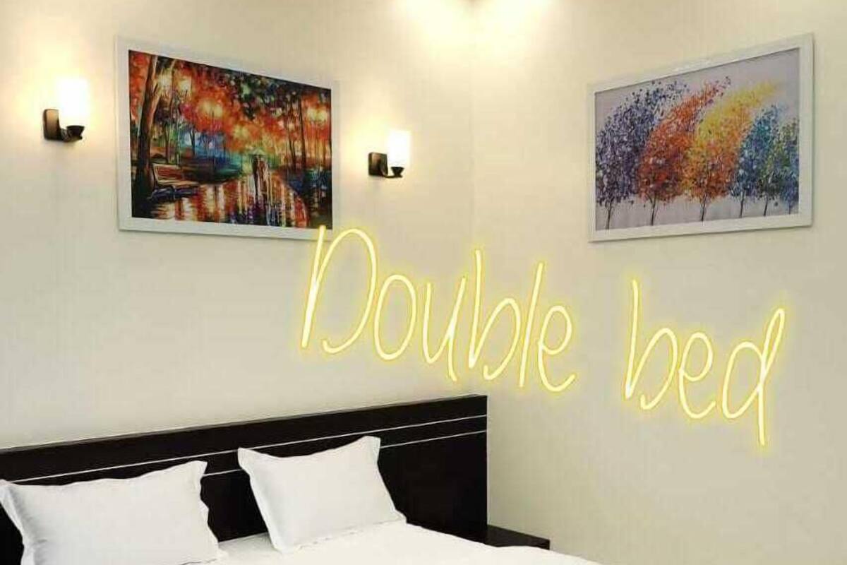 A double bed with white linens, two wall-mounted light fixtures, and two framed paintings above the bed.