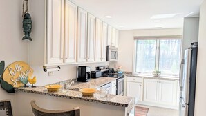 A modern kitchen with white cabinets, granite countertops, and stainless steel appliances. There are two chairs and a small island with a bowl and a coffee maker.