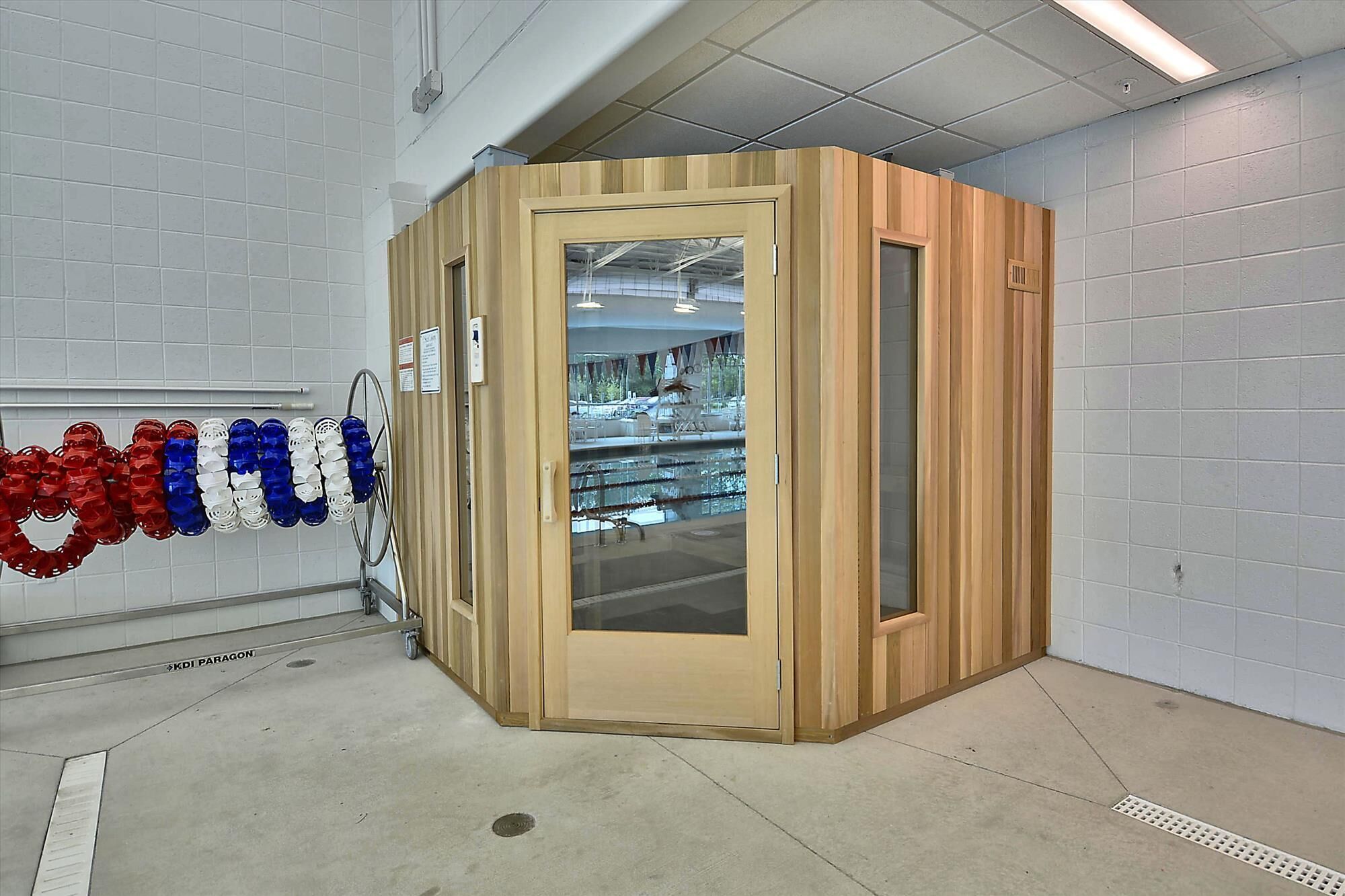 A compact, wooden room with a glass door, white tiled walls, and a concrete floor.