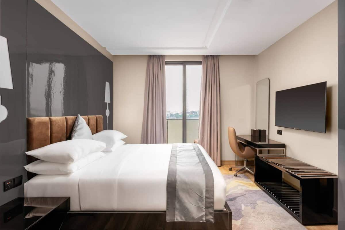A modern hotel room with a large bed, a desk, a chair, a TV, and a view of the city through the window.