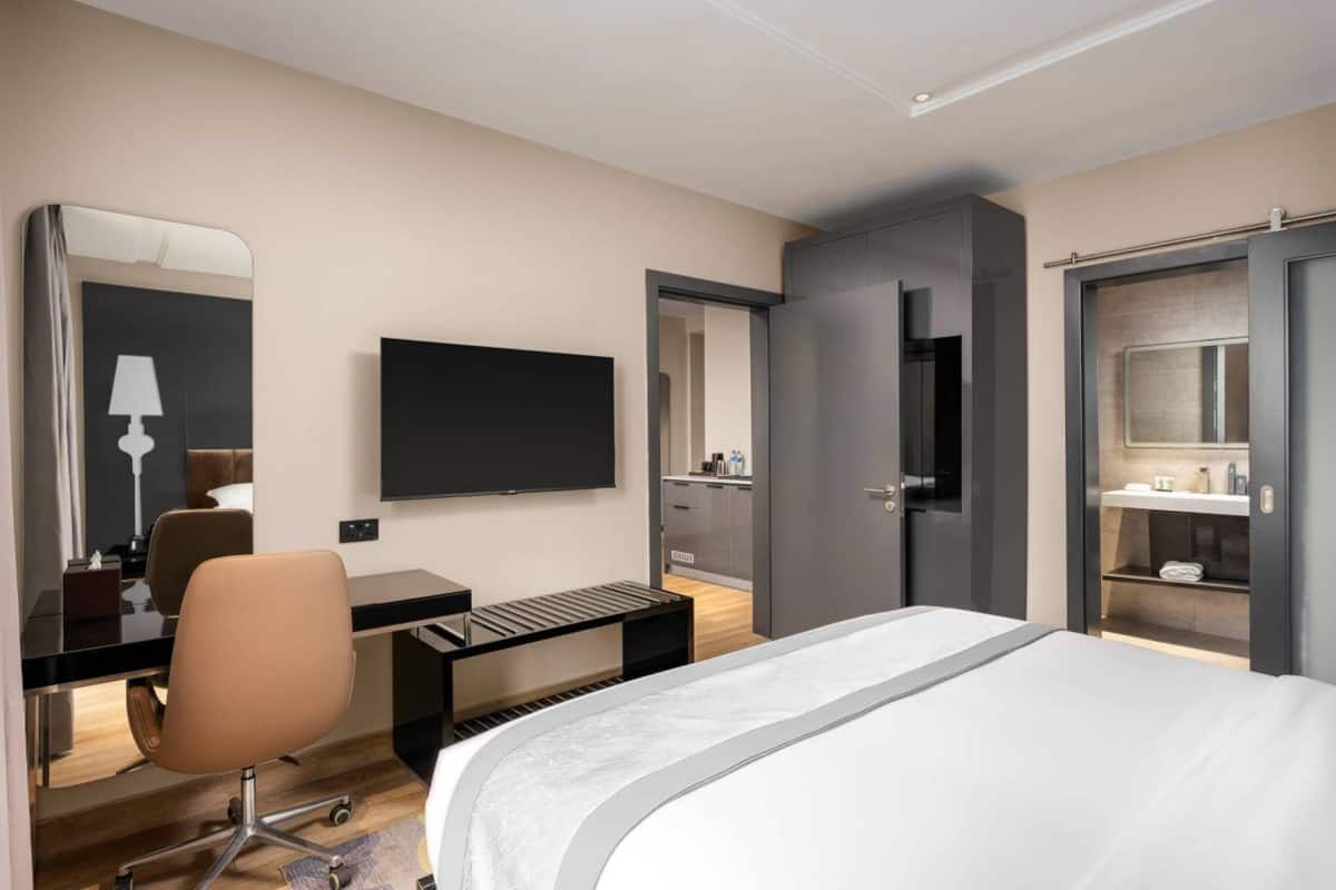 A modern hotel room with a large bed, a desk with a chair, a TV, a mirror, and a wardrobe.