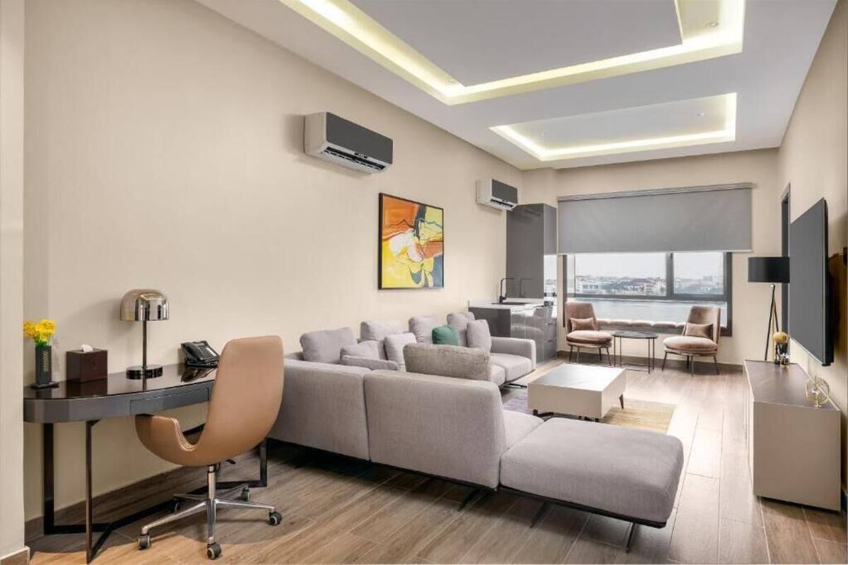 A modern living room with a sectional sofa, a desk with a lamp, a flat-screen TV, and a large window with a city view.
