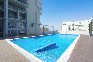 A rooftop swimming pool with a tiled deck, surrounded by modern buildings with balconies. - Bloomfields Studio in Oasis 1 - Masdar (Abu Dhabi)