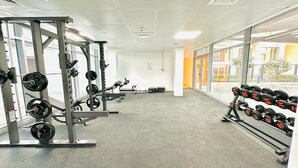 A gym with weightlifting equipment, including barbells and dumbbells, and large windows offering a view of the outside. - Bloomfields Grandeur STU in Oasis Masdar (Abu Dhabi)